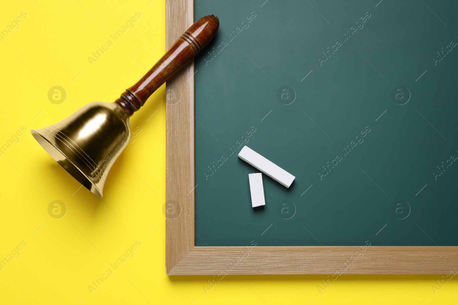 Photo of Golden bell, green board and pieces of chalk on yellow background, flat lay. School days