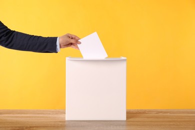 Photo of Woman putting her vote into ballot box on wooden table against orange background, closeup