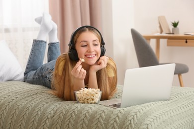 Teenage girl with headphones eating popcorn while using laptop on bed at home