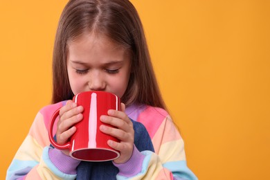 Photo of Cute girl drinking beverage from red ceramic mug on orange background, space for text