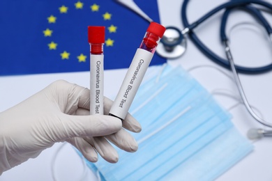 Photo of Doctor holding sample tubes with labels Coronavirus Blood Test above medical items and European Union flag, closeup