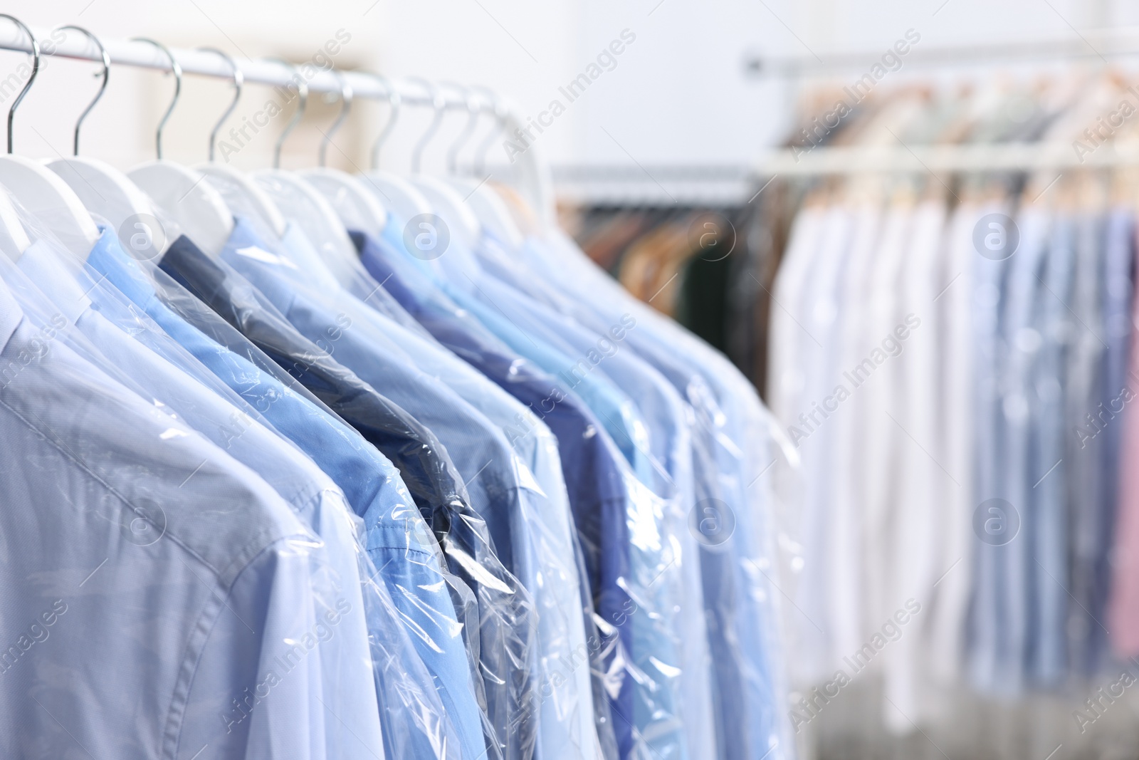 Photo of Dry-cleaning service. Many different clothes in plastic bags hanging on rack indoors, closeup