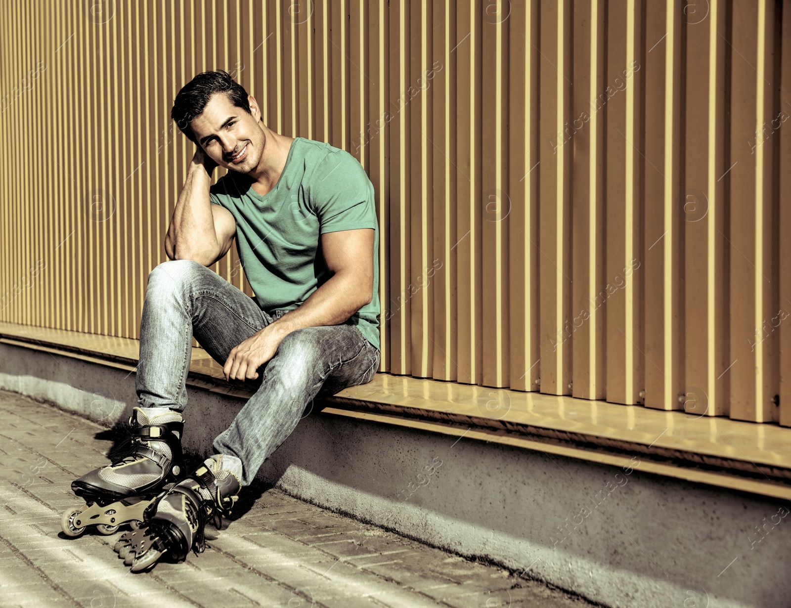 Image of Handsome young man with inline roller skates sitting near yellow building