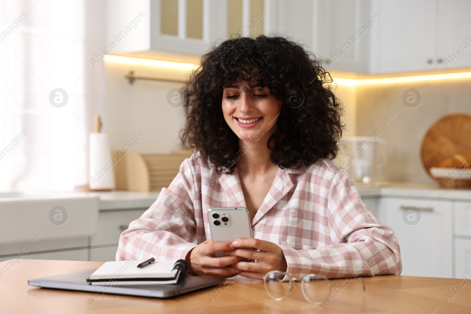 Photo of Beautiful young woman in stylish pyjama using smartphone at wooden table in kitchen