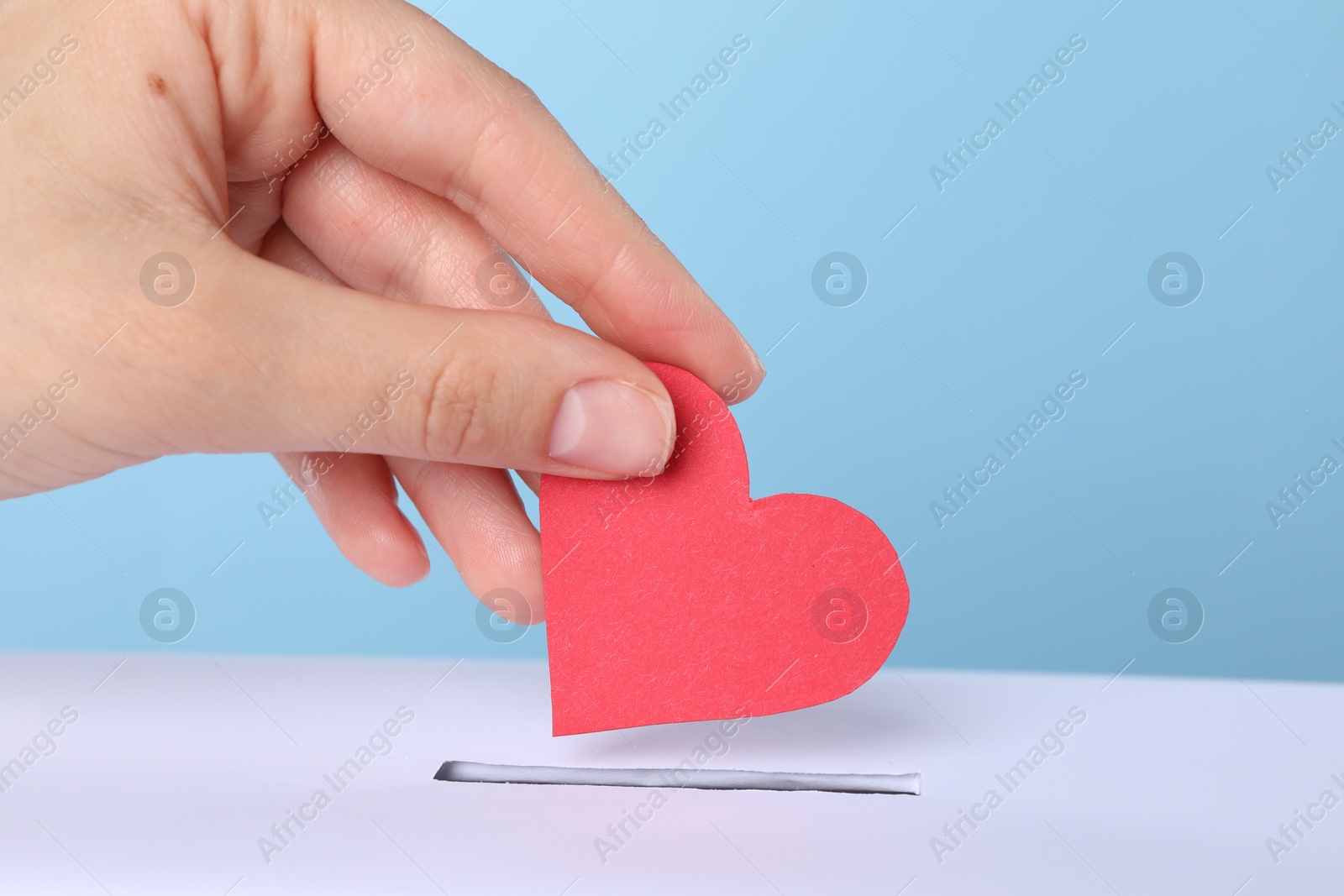 Photo of Woman putting red heart into slot of donation box against light blue background, closeup