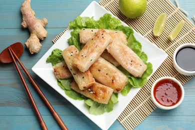 Photo of Tasty fried spring rolls, lettuce, lime and sauces on light blue wooden table, flat lay