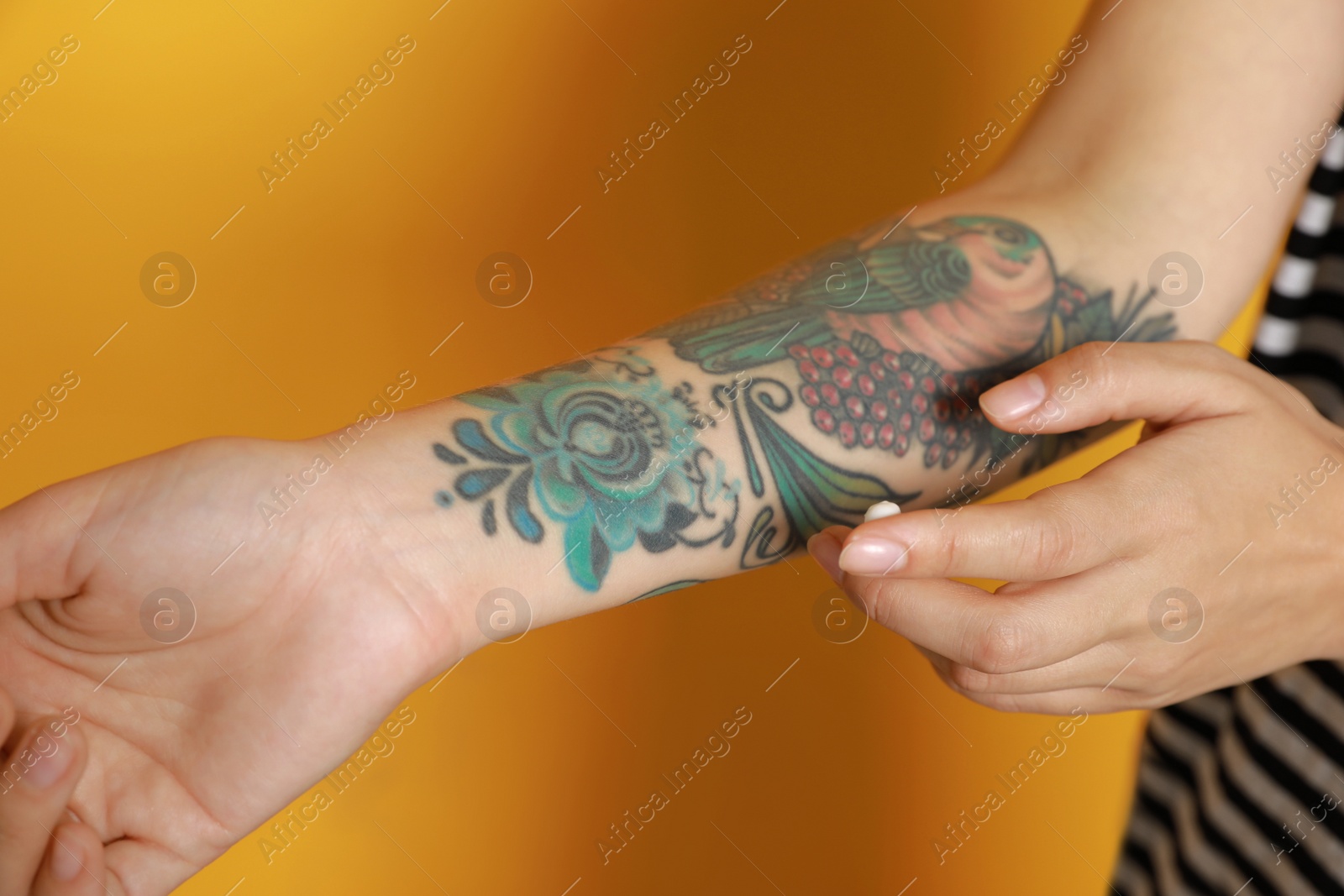 Photo of Woman applying cream on her arm with tattoos against yellow background, closeup