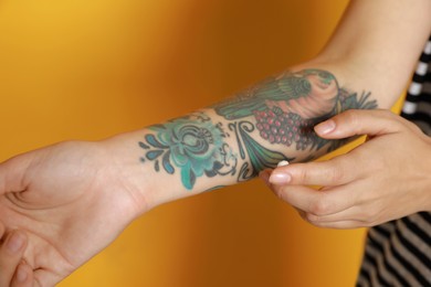 Woman applying cream on her arm with tattoos against yellow background, closeup