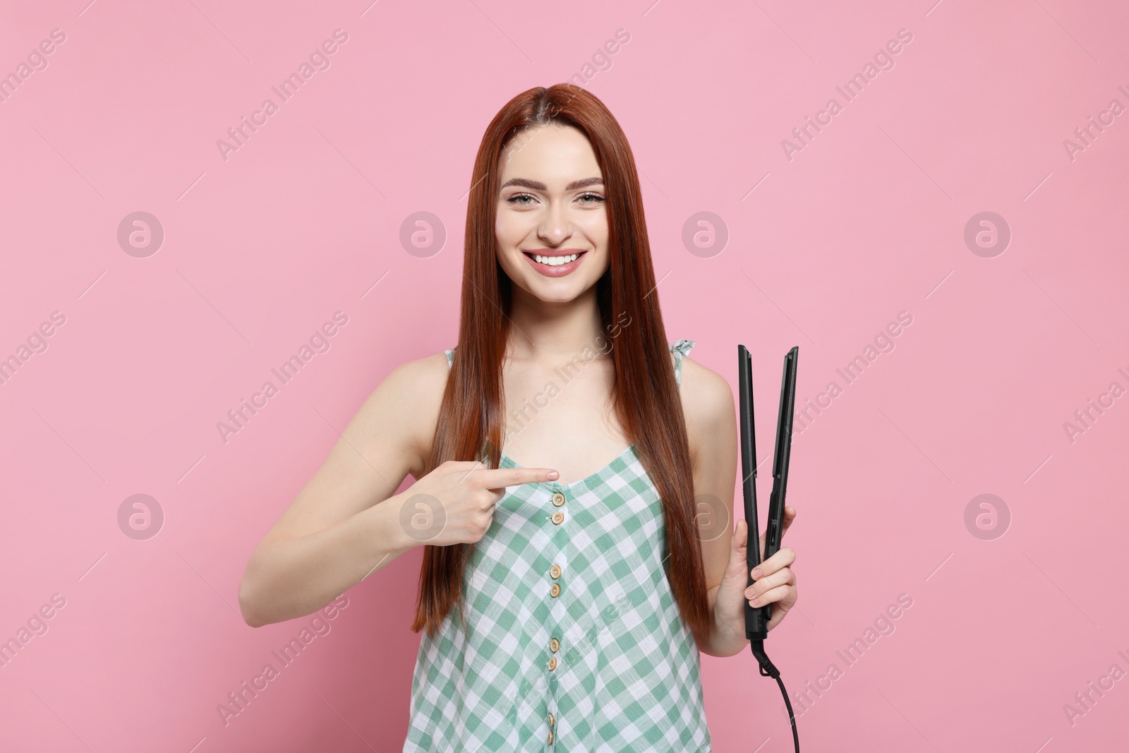 Photo of Beautiful woman pointing at hair iron on pink background