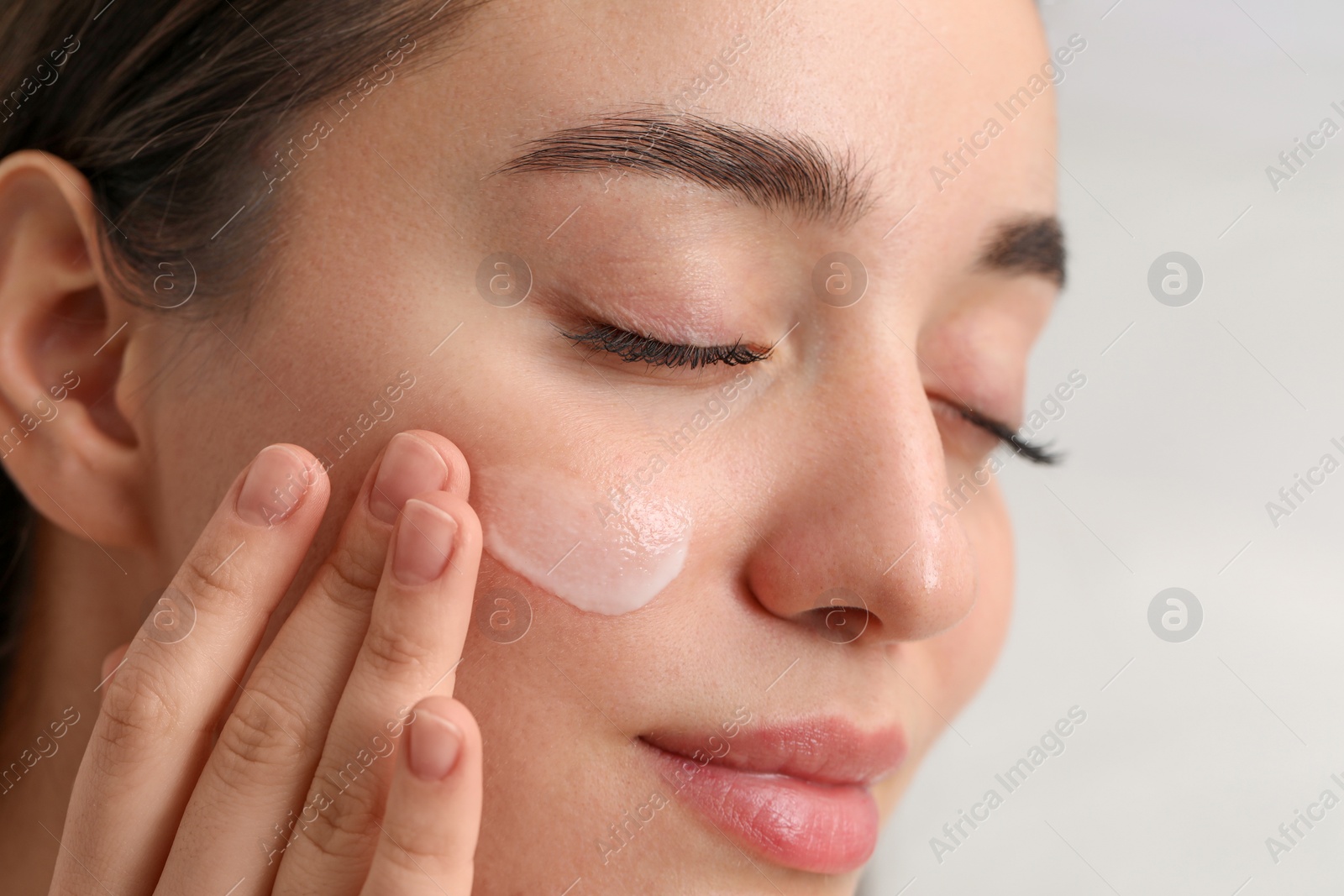 Photo of Young woman with dry skin applying cream onto her face on blurred background, closeup