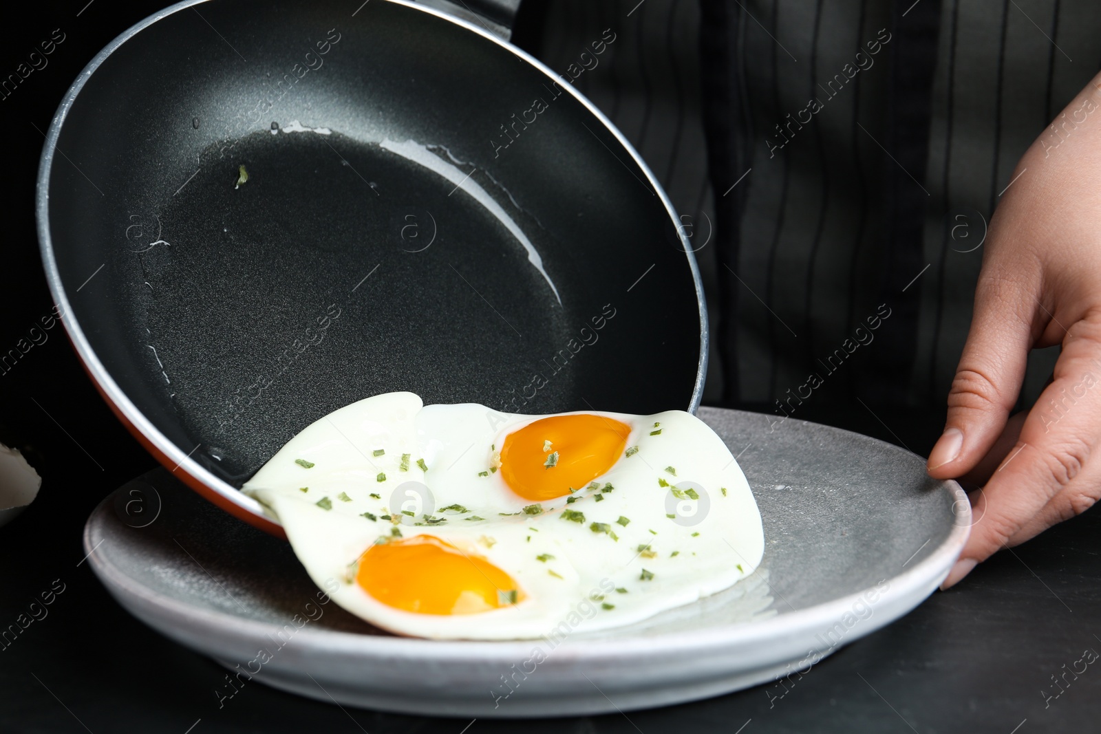 Photo of Woman putting tasty cooked eggs onto plate from frying pan at black table, closeup