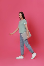 Young woman with modern laptop walking on pink background