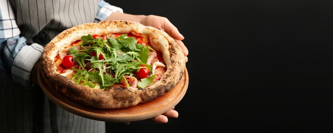 Image of Woman holding tasty pizza with meat and arugula on black background, closeup view with space for text. Banner design