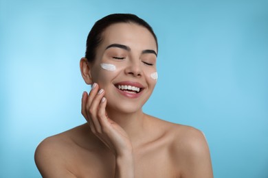 Young woman applying facial cream on light blue background