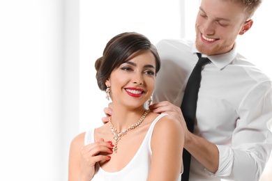 Man putting elegant jewelry on beautiful woman against light background