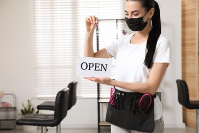 Photo of Professional stylist with protective mask holding Open sign in salon, space for text. Hairdressing services during Coronavirus quarantine