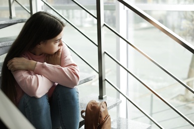 Photo of Upset teenage girl with backpack sitting on stairs indoors. Space for text