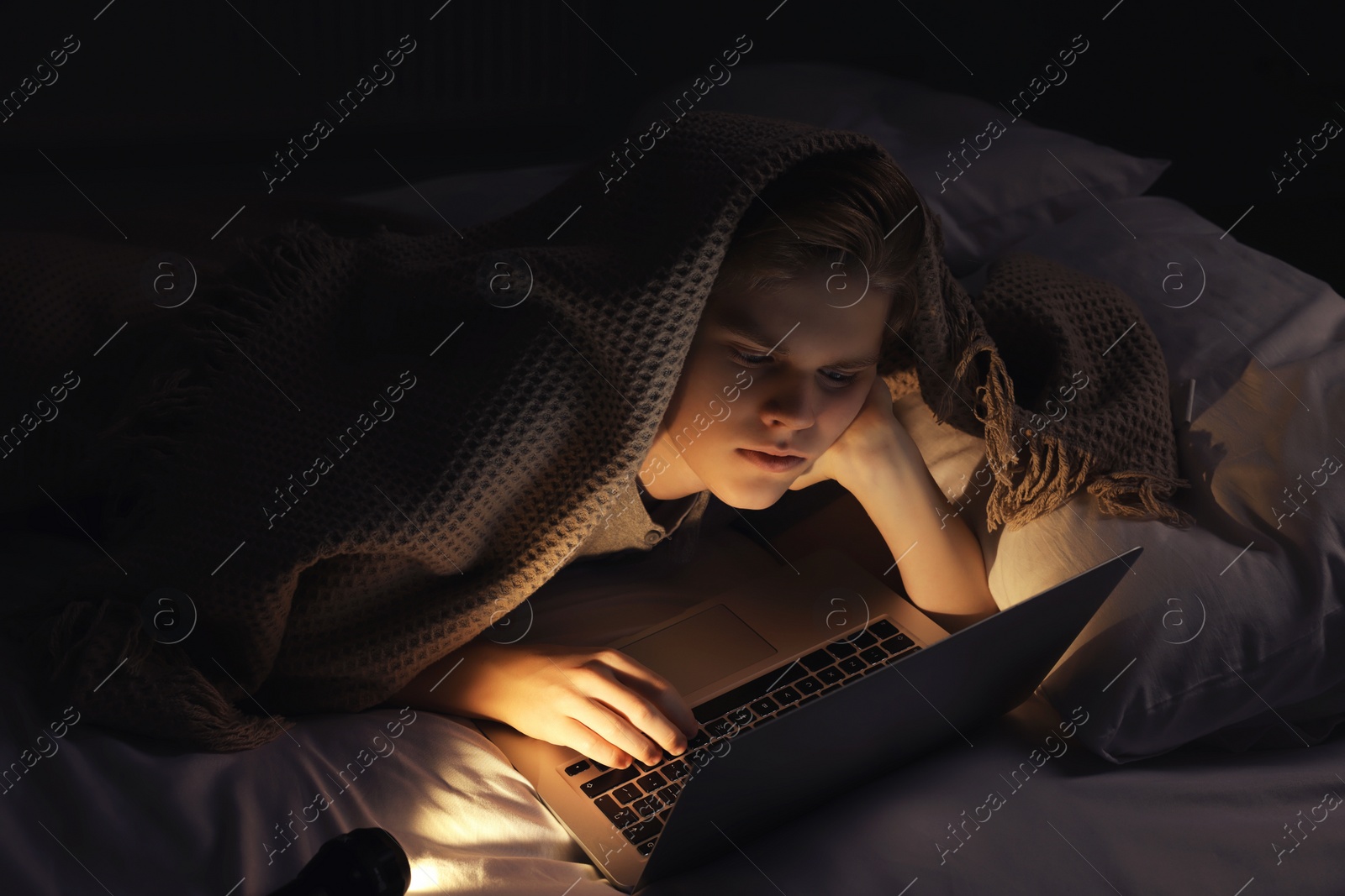 Photo of Teenage boy using laptop under blanket on bed at night. Internet addiction