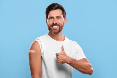 Photo of Man with sticking plaster on arm after vaccination showing thumbs up against light blue background