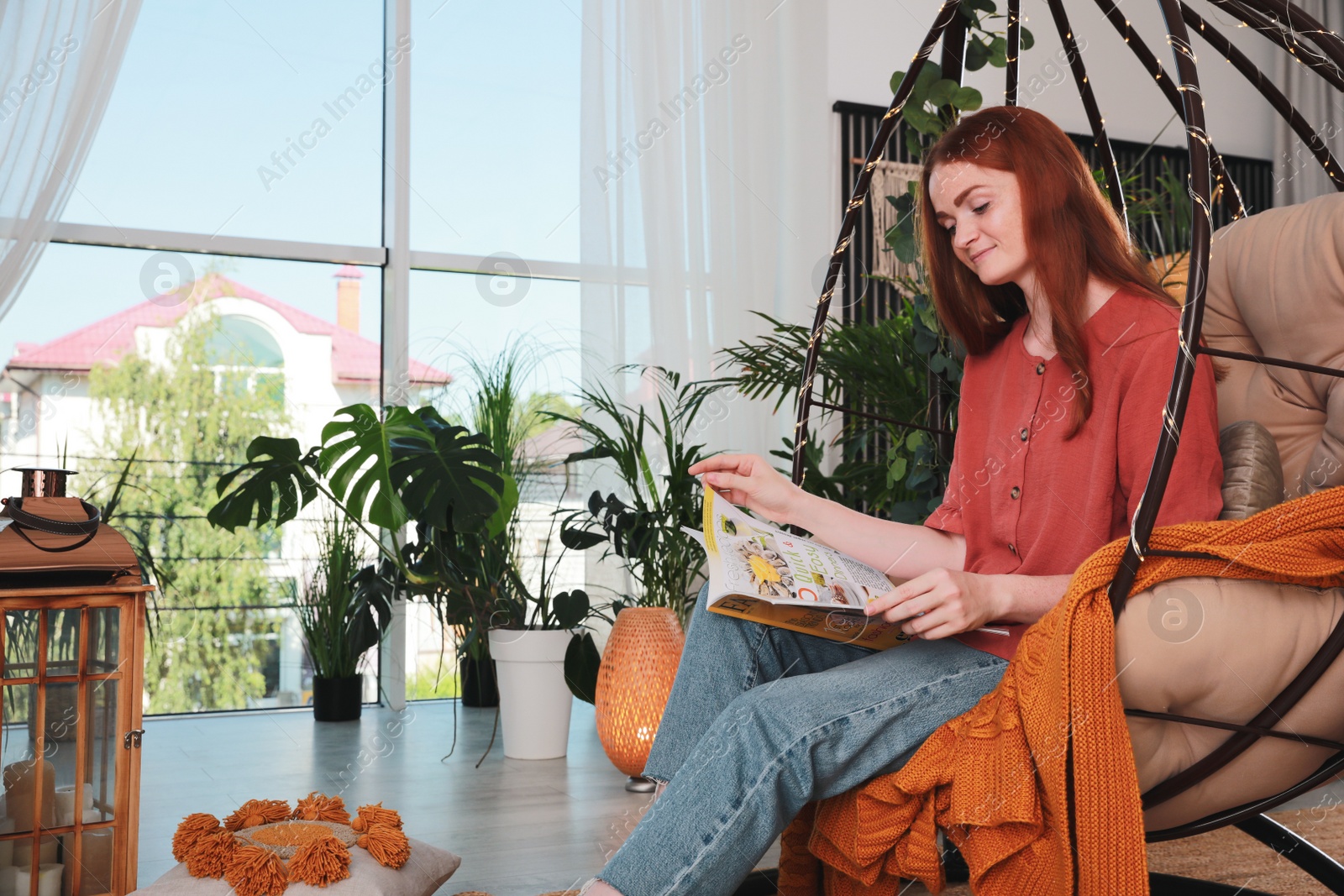 Photo of Happy young woman reading magazine in egg chair at indoor terrace