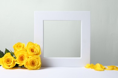 Photo of Beautiful presentation for product. Frame and yellow roses on white table against light grey background, space for text