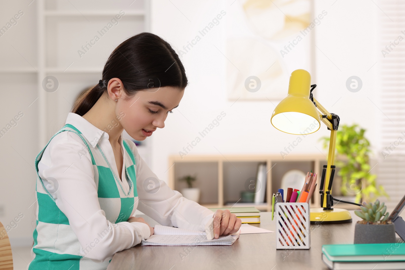 Photo of Teenage girl erasing mistake in her notebook at wooden desk indoors