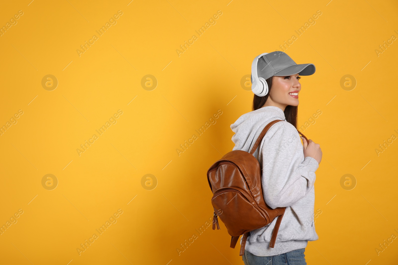Photo of Beautiful young woman with stylish leather backpack and headphones on yellow background. Space for text