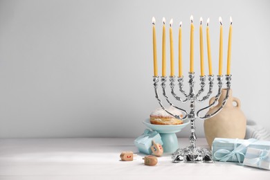 Photo of Hanukkah celebration. Menorah with burning candles, dreidels and gift boxes on white table, space for text