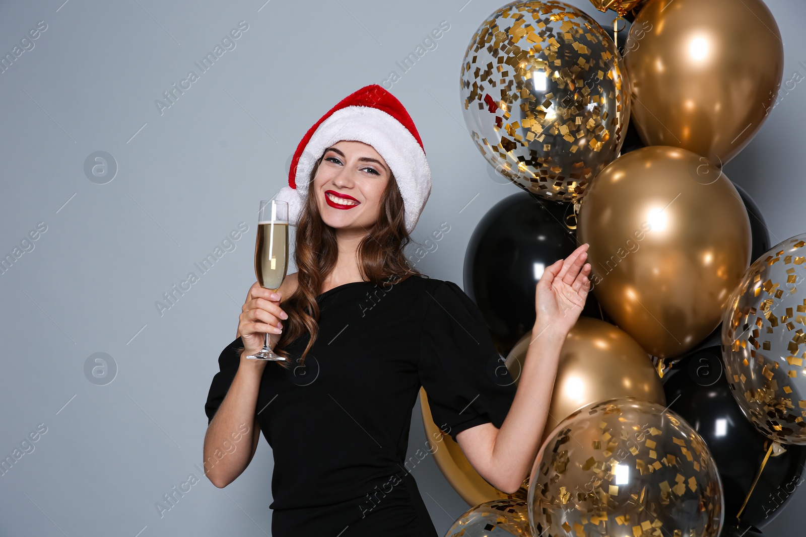 Photo of Happy woman in Santa hat with glass of champagne near air balloons on grey background. Christmas party