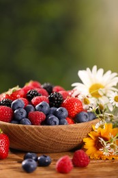 Photo of Bowl with different fresh ripe berries and beautiful flowers on wooden table outdoors