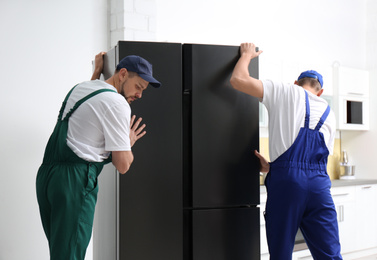 Professional workers carrying modern refrigerator in kitchen