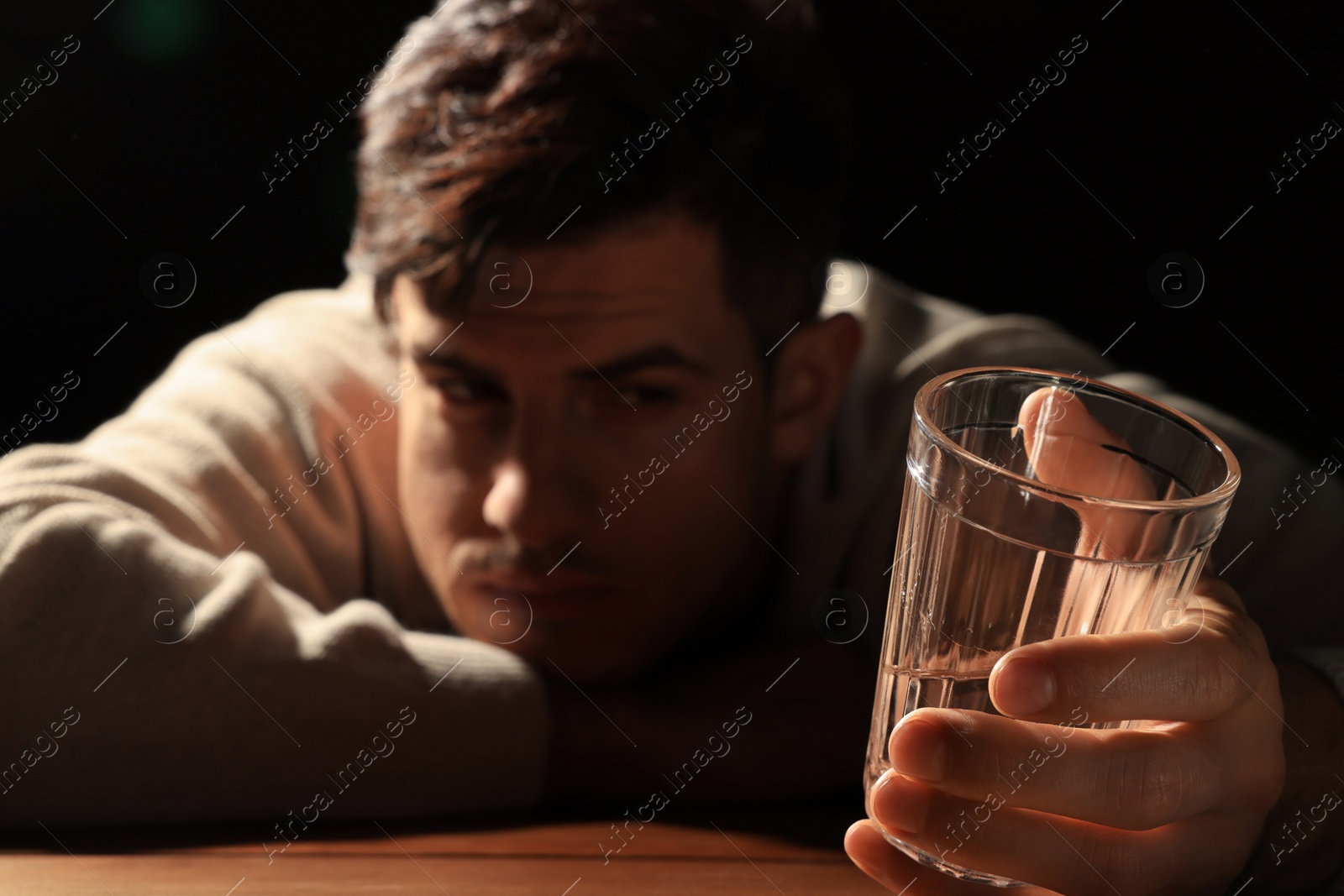 Photo of Addicted man at wooden table against black background, focus on glass of alcoholic drink