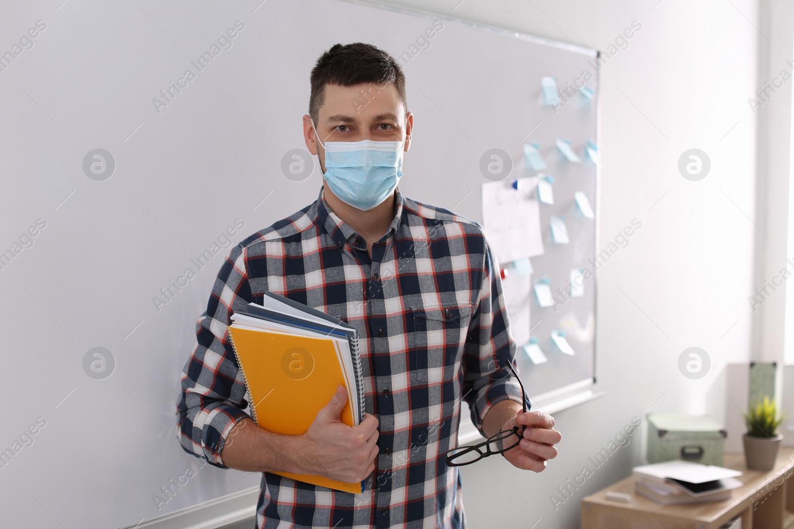 Photo of Teacher with protective mask, glasses and copybooks near board in classroom. Reopening after Covid-19 quarantine