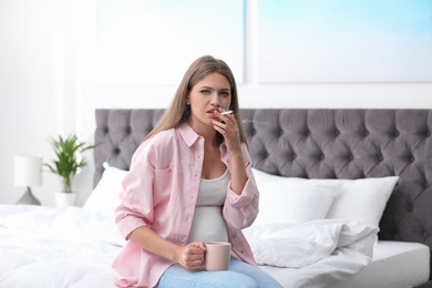 Photo of Young pregnant woman with cup of coffee smoking cigarette in bedroom. Harm to unborn baby