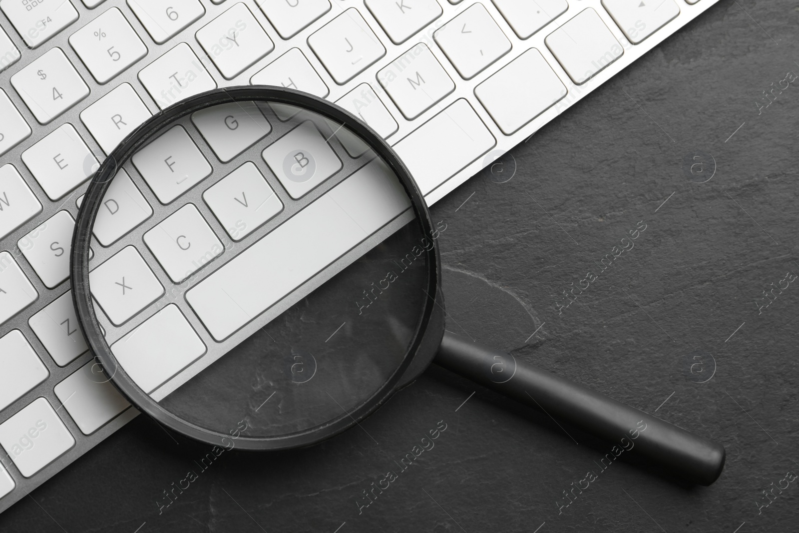 Photo of Magnifier glass and keyboard on black slate background, flat lay. Find keywords concept