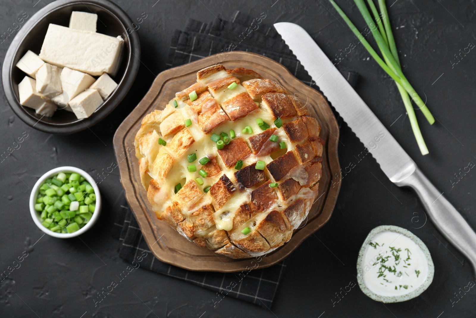 Photo of Freshly baked bread with tofu cheese, green onions, sauce and knife on black table, flat lay