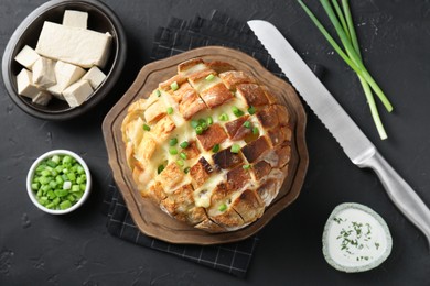 Photo of Freshly baked bread with tofu cheese, green onions, sauce and knife on black table, flat lay