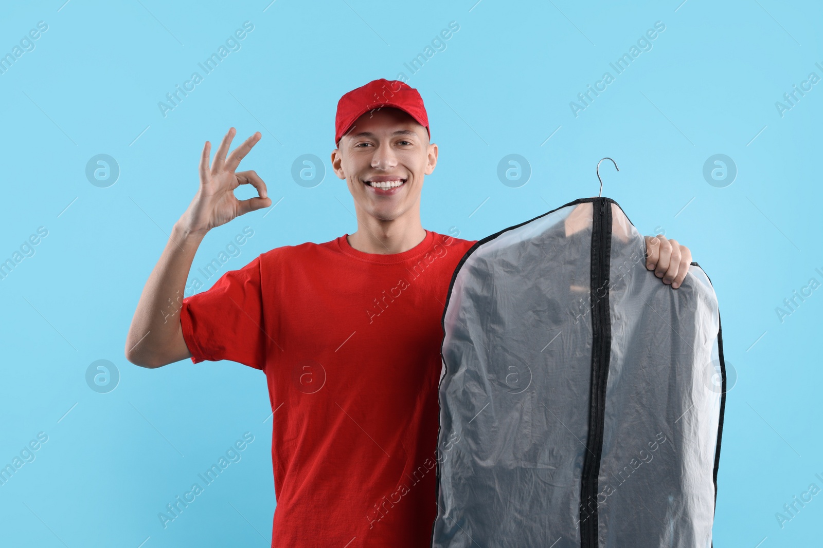 Photo of Dry-cleaning delivery. Happy courier holding garment cover with clothes and showing OK gesture on light blue background