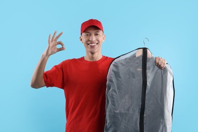 Photo of Dry-cleaning delivery. Happy courier holding garment cover with clothes and showing OK gesture on light blue background