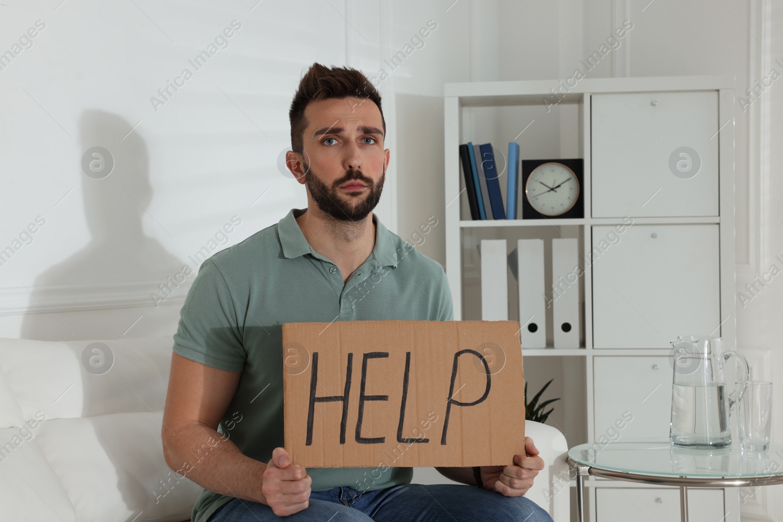 Photo of Unhappy man with HELP sign on sofa indoors