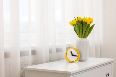 Photo of Spring interior. Beautiful yellow tulips and clock on white chest of drawers near window, space for text