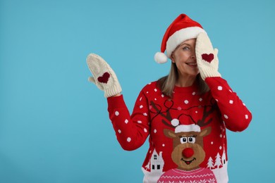 Senior woman in Christmas sweater, Santa hat and knitted mittens on light blue background. Space for text