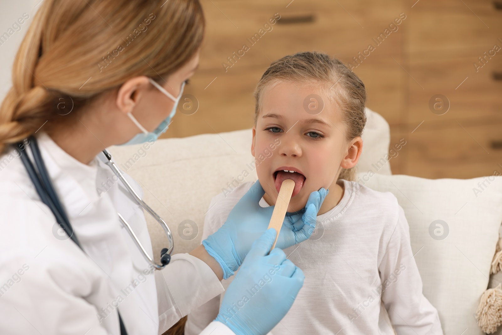 Photo of Doctor in medical mask examining girl`s oral cavity with tongue depressor indoors