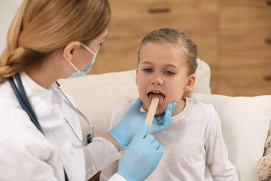 Photo of Doctor in medical mask examining girl`s oral cavity with tongue depressor indoors