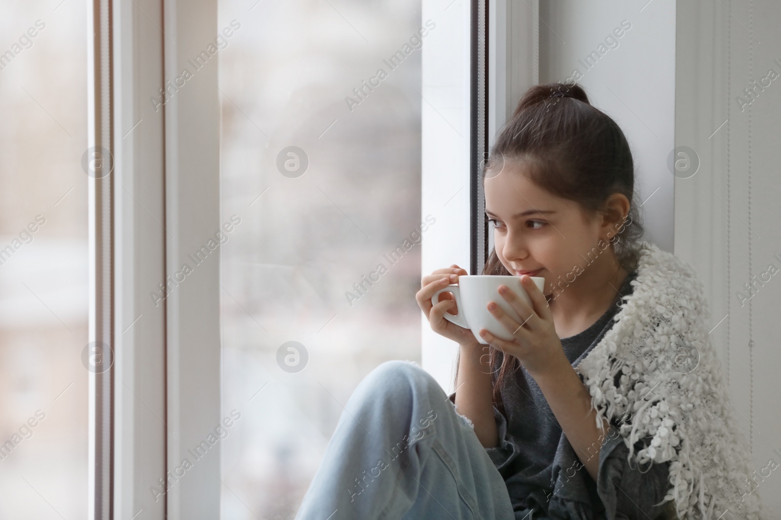 Photo of Cute little girl with cup of drink resting near window at home. Space for text