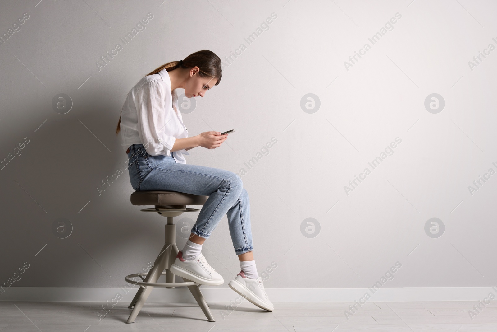 Photo of Woman with bad posture using smartphone while sitting on stool near light grey wall indoors, space for text