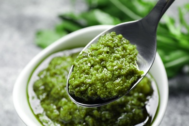 Photo of Spoon of tasty arugula pesto near bowl with sauce, closeup