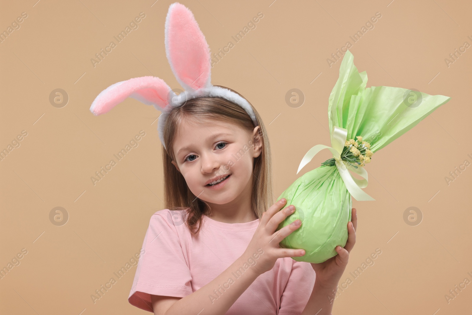 Photo of Easter celebration. Cute girl with bunny ears holding wrapped gift on beige background