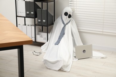 Photo of Overworked ghost. Man in white sheet with laptop on floor in office
