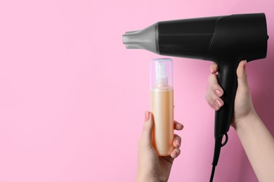 Woman holding spray bottle with thermal protection and hairdryer on light pink background, closeup. Space for text